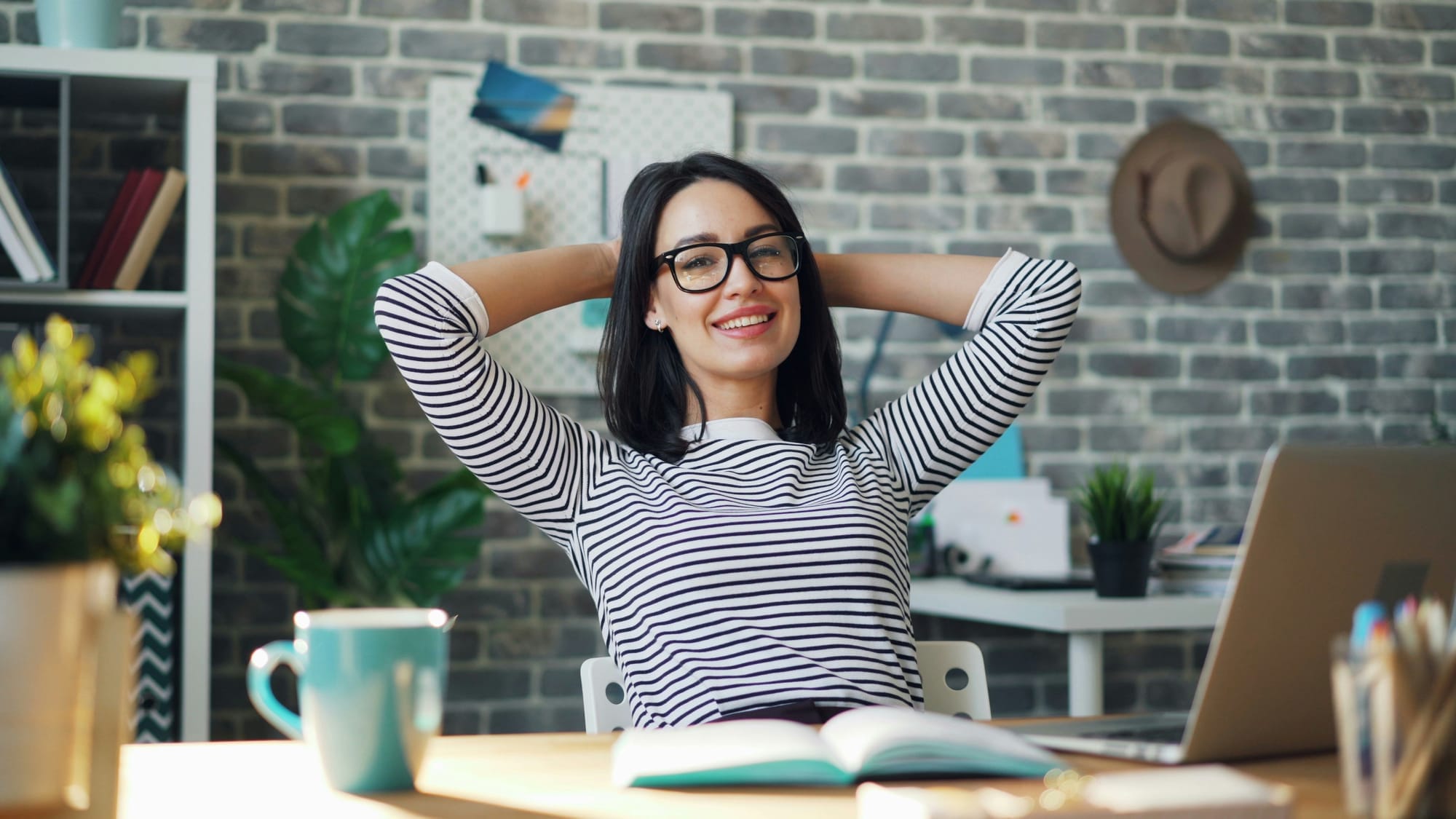 A dark haired woman putting her hands behind her head and smiling. She is sitting at a desk with an open book and a turqoiuse mug.