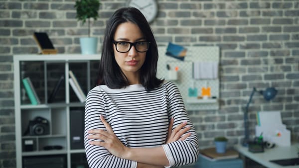 A dark-haired woman standing in front of a brick wall with crossed arms, wearing a black and white striped shrit